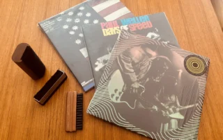 Three vinyl records with three different record cleaning brushes arranged on a table showing vinyl care tools and albums